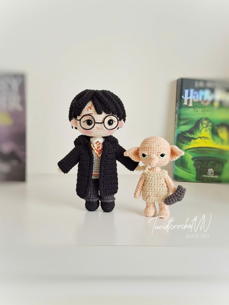 May include: Crocheted Harry Potter and Dobby dolls. Harry wears a black coat, glasses, and has a lightning bolt scar. Dobby is beige and holds a gray item. The text "TuoidtcrochetN SINCE 2023" is visible.