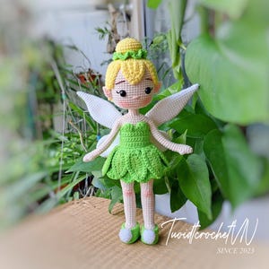 May include: A handmade crochet doll of a fairy with yellow hair, a green dress, and white wings. The doll has a sweet expression and is wearing green shoes. The doll is standing on a woven surface, with greenery in the background.