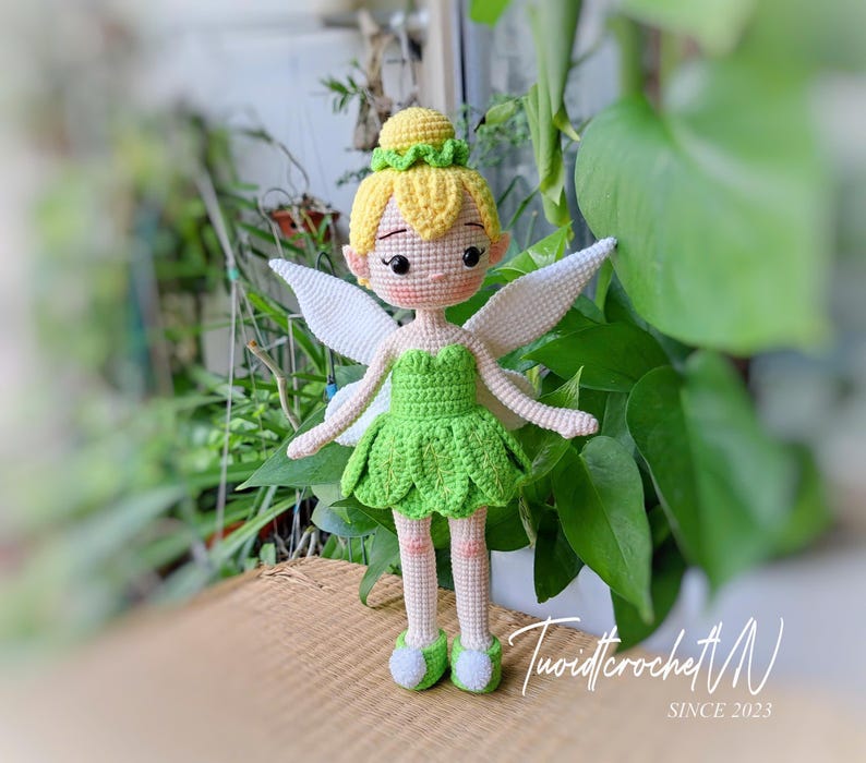 May include: A handmade crochet doll of a fairy with yellow hair, a green dress, and white wings. The doll has a sweet expression and is wearing green shoes. The doll is standing on a woven surface, with greenery in the background.