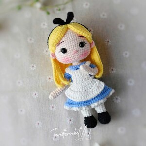Puede incluir: Mu&ntilde;eca de ganchillo hecha a mano de Alicia en el Pa&iacute;s de las Maravillas. La mu&ntilde;eca tiene cabello amarillo, un lazo negro y viste un vestido blanco con detalles azules. La cara est&aacute; detallada con ojos negros y una nariz rosa.