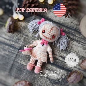 Puede incluir: Una muñeca de ganchillo con cabello de hilo gris en coletas, ojos de botón rojos y detalles de boca cosidos. La muñeca viste un vestido blanco con detalles en rosa y sostiene un pequeño juguete a juego. El texto dice PDF PATTERN y 16CM.