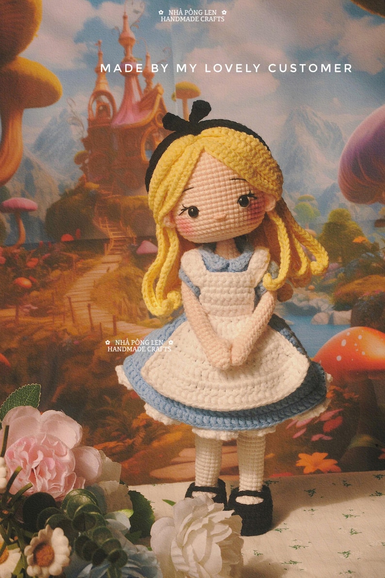 May include: Crocheted doll of a character from Alice in Wonderland, wearing a blue and white dress, black shoes, and a black bow. The doll has blonde hair and rosy cheeks. The background features a whimsical landscape with a castle and mushrooms. Text reads "MADE BY MY LOVELY CUSTOMER".