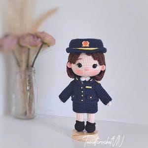 May include: A handcrafted crochet doll wearing a dark navy blue uniform consisting of a jacket, skirt, and matching peaked cap with gold accents. The doll has brown hair, rosy cheeks, and stands on a small circular wooden base. The text Tuoidcrochet N SINCE 2023 is visible.
