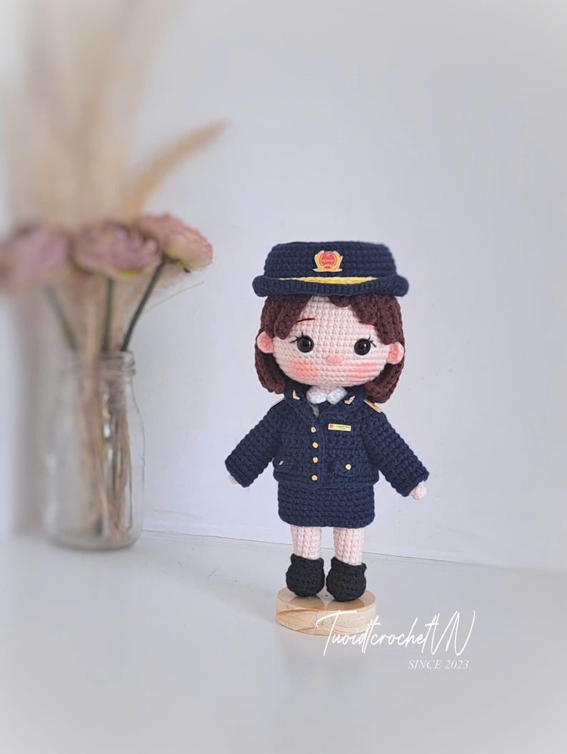 May include: A handcrafted crochet doll wearing a dark navy blue uniform consisting of a jacket, skirt, and matching peaked cap with gold accents. The doll has brown hair, rosy cheeks, and stands on a small circular wooden base. The text Tuoidcrochet N SINCE 2023 is visible.