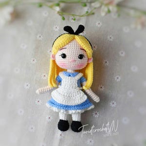 Puede incluir: Mu&ntilde;eca de ganchillo hecha a mano de Alicia en el pa&iacute;s de las maravillas. La mu&ntilde;eca tiene cabello amarillo, un lazo negro y viste un vestido azul y blanco. La cara est&aacute; detallada con ojos negros y boca rosa. La mu&ntilde;eca mide aproximadamente 15 cm de alto.