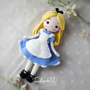May include: A handmade crochet doll depicting Alice in Wonderland. The doll features a blue dress with a white apron, yellow hair, and black shoes. The doll is lying down on a gray surface with white flowers. The text "TuoidtcrochetVN SINCE 2023" is visible.