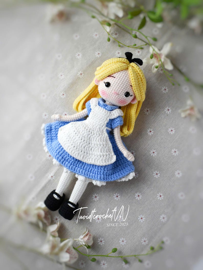 May include: A handmade crochet doll depicting Alice in Wonderland. The doll features a blue dress with a white apron, yellow hair, and black shoes. The doll is lying down on a gray surface with white flowers. The text "TuoidtcrochetVN SINCE 2023" is visible.