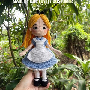 May include: A handmade crochet doll of Alice in Wonderland, featuring a blue dress with a white apron, yellow hair, and black shoes. The doll is held in front of a lush green background. Text on the image reads "MADE BY OUR LOVELY CUSTOMER".