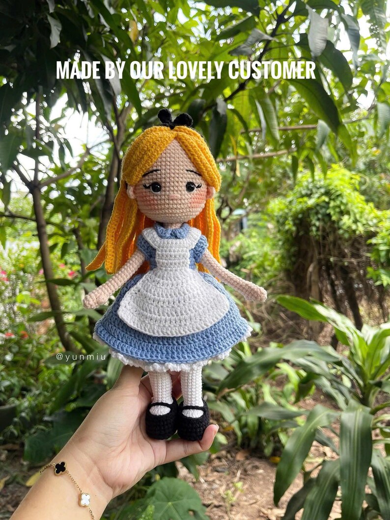 May include: A handmade crochet doll of Alice in Wonderland, featuring a blue dress with a white apron, yellow hair, and black shoes. The doll is held in front of a lush green background. Text on the image reads "MADE BY OUR LOVELY CUSTOMER".