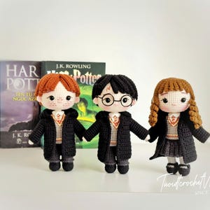 May include: Three handmade crochet dolls inspired by the Harry Potter series. Each doll wears a black robe and a school uniform. The dolls have different hair colors and styles. Books are in the background.