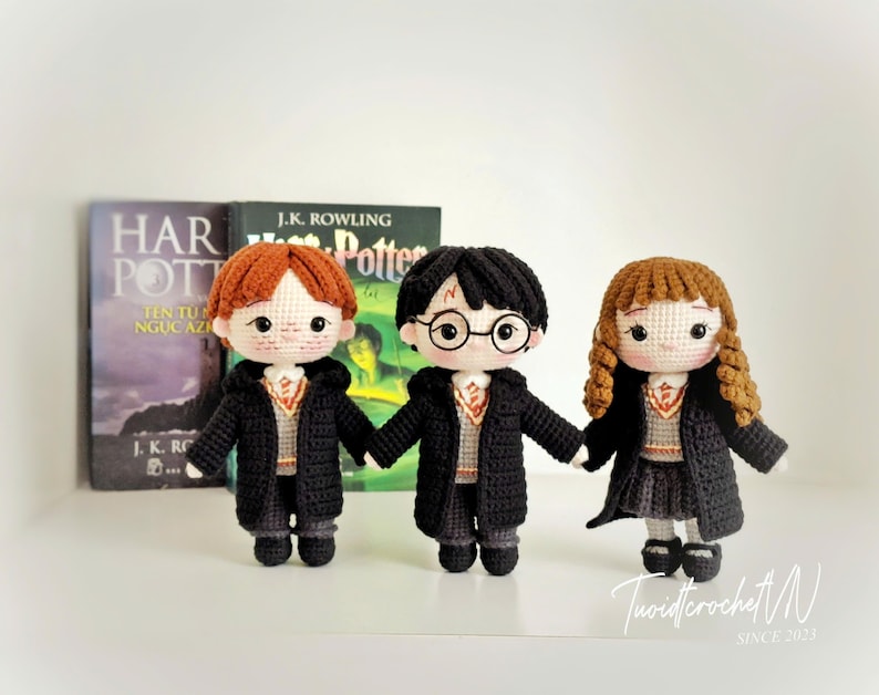 May include: Three handmade crochet dolls inspired by the Harry Potter series. Each doll wears a black robe and a school uniform. The dolls have different hair colors and styles. Books are in the background.