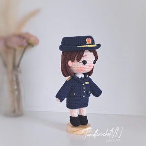 May include: A crocheted doll wearing a dark blue uniform, a matching peaked cap with a yellow emblem, and black shoes. The doll has brown hair, pink cheeks, and stands on a circular wooden base. The text "Tuoidcrochet N SINCE 2023" is visible.