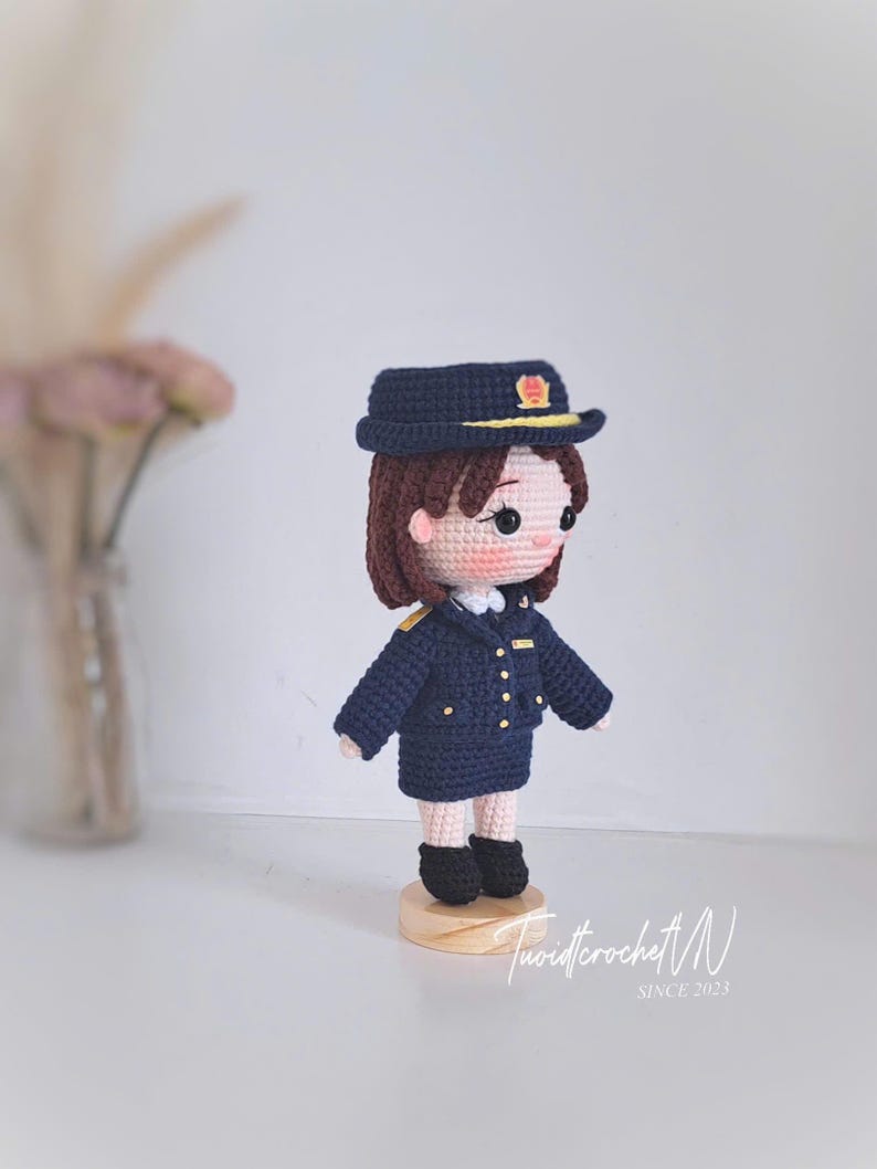 May include: A crocheted doll wearing a dark blue uniform, a matching peaked cap with a yellow emblem, and black shoes. The doll has brown hair, pink cheeks, and stands on a circular wooden base. The text "Tuoidcrochet N SINCE 2023" is visible.