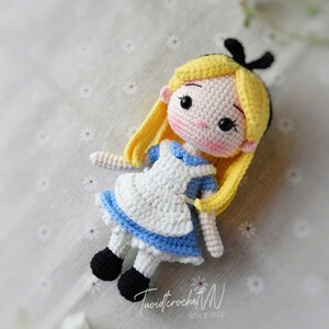 Puede incluir: Mu&ntilde;eca de ganchillo de Alicia en el pa&iacute;s de las maravillas. La mu&ntilde;eca tiene pelo amarillo, un vestido azul y blanco y zapatos negros. La mu&ntilde;eca est&aacute; sobre una superficie estampada. El texto "TwistcrochetM SINCE 2023" es visible.