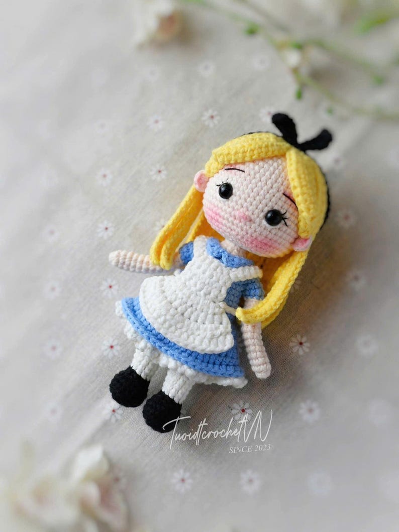 Puede incluir: Mu&ntilde;eca de ganchillo de Alicia en el pa&iacute;s de las maravillas. La mu&ntilde;eca tiene pelo amarillo, un vestido azul y blanco y zapatos negros. La mu&ntilde;eca est&aacute; sobre una superficie estampada. El texto "TwistcrochetM SINCE 2023" es visible.