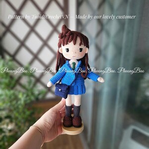 Pode incluir: Boneca de croch&ecirc; feita &agrave; m&atilde;o de uma menina com uniforme escolar azul, com blazer, saia plissada e uma pequena bolsa. A boneca tem cabelo castanho e est&aacute; sobre uma base de madeira. O texto "Pattern by TuoidtCrochetVN - Made by our lovely customer" &eacute; vis&iacute;vel.