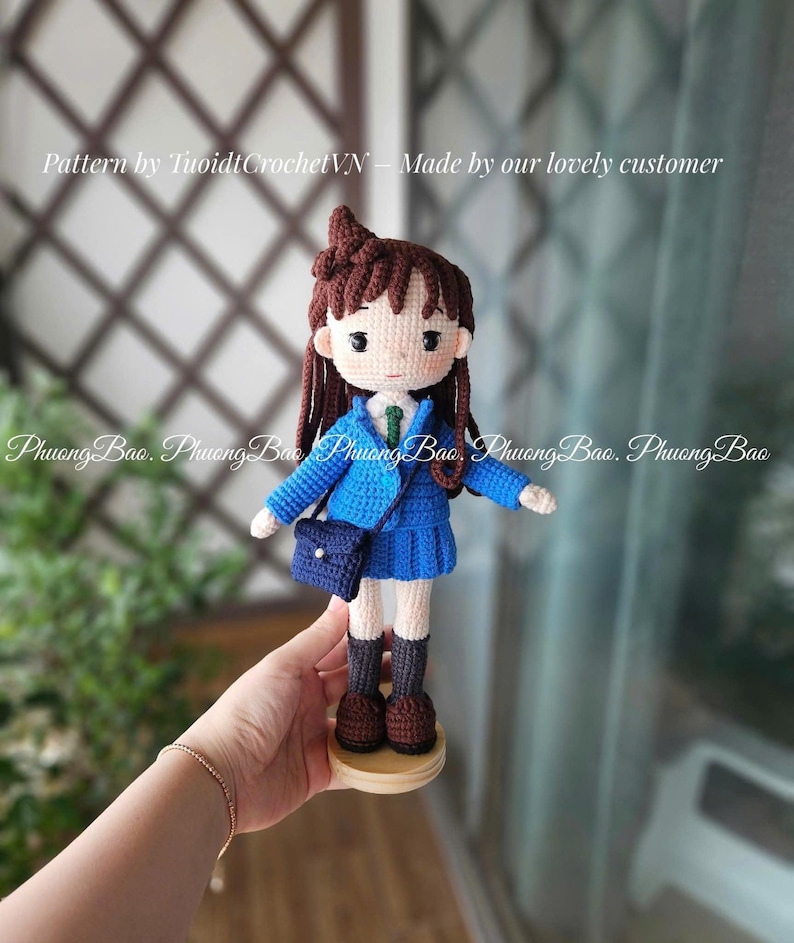 Pode incluir: Boneca de croch&ecirc; feita &agrave; m&atilde;o de uma menina com uniforme escolar azul, com blazer, saia plissada e uma pequena bolsa. A boneca tem cabelo castanho e est&aacute; sobre uma base de madeira. O texto "Pattern by TuoidtCrochetVN - Made by our lovely customer" &eacute; vis&iacute;vel.
