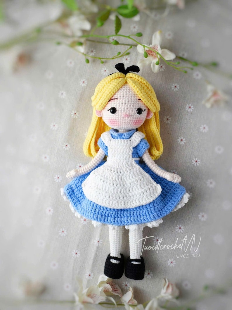 May include: A handmade crochet doll of Alice in Wonderland. The doll has yellow hair, a blue dress with a white apron, and black shoes. The doll is made with a crochet stitch and has a sweet, innocent expression.