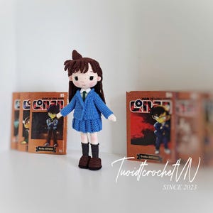 Pode incluir: Boneca de croch&ecirc; feita &agrave; m&atilde;o de um personagem da s&eacute;rie Detetive Conan. A boneca usa um blazer e saia azuis, com cabelo castanho e botas pretas. Atr&aacute;s, v&aacute;rios mang&aacute;s de Detetive Conan.