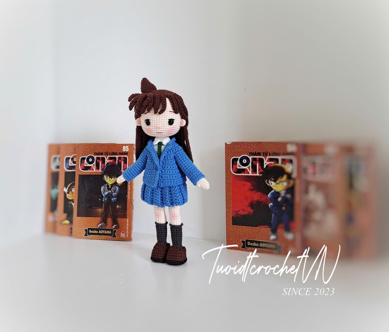 Pode incluir: Boneca de croch&ecirc; feita &agrave; m&atilde;o de um personagem da s&eacute;rie Detetive Conan. A boneca usa um blazer e saia azuis, com cabelo castanho e botas pretas. Atr&aacute;s, v&aacute;rios mang&aacute;s de Detetive Conan.
