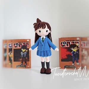Pode incluir: Boneca de croch&ecirc; feita &agrave; m&atilde;o de um personagem com uniforme escolar azul, cabelo castanho e sapatos pretos. A boneca est&aacute; em frente a v&aacute;rias hist&oacute;rias em quadrinhos com a palavra "Conan" na capa. &Eacute; um item feito &agrave; m&atilde;o.