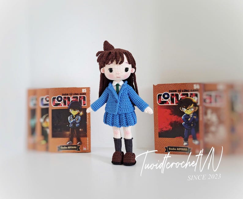Pode incluir: Boneca de croch&ecirc; feita &agrave; m&atilde;o de um personagem com uniforme escolar azul, cabelo castanho e sapatos pretos. A boneca est&aacute; em frente a v&aacute;rias hist&oacute;rias em quadrinhos com a palavra "Conan" na capa. &Eacute; um item feito &agrave; m&atilde;o.