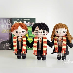 May include: Three handmade crochet dolls of characters from the Harry Potter series. The dolls have black, red, and brown hair, and wear black robes and striped scarves. A book with the title "Harry Potter" is in the background.