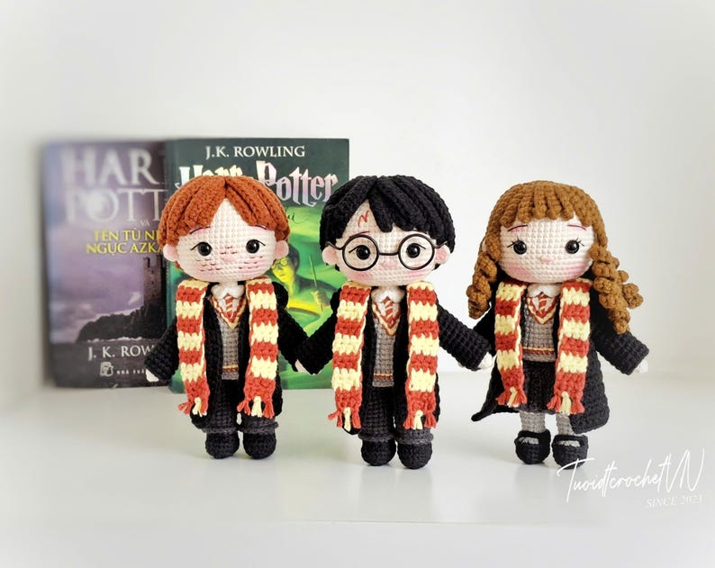 May include: Three handmade crochet dolls of characters from the Harry Potter series. The dolls have black, red, and brown hair, and wear black robes and striped scarves. A book with the title "Harry Potter" is in the background.