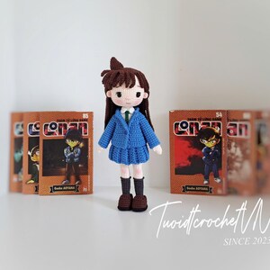 Pode incluir: Boneca de croch&ecirc; feita &agrave; m&atilde;o de um personagem da s&eacute;rie de mang&aacute; Conan, usando um uniforme escolar azul. A boneca tem cabelo castanho e est&aacute; em frente a v&aacute;rios livros de mang&aacute;. A boneca tem cerca de 15 cm de altura.