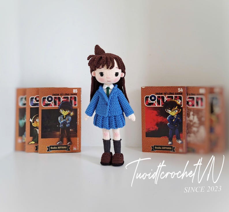 Pode incluir: Boneca de croch&ecirc; feita &agrave; m&atilde;o de um personagem da s&eacute;rie de mang&aacute; Conan, usando um uniforme escolar azul. A boneca tem cabelo castanho e est&aacute; em frente a v&aacute;rios livros de mang&aacute;. A boneca tem cerca de 15 cm de altura.