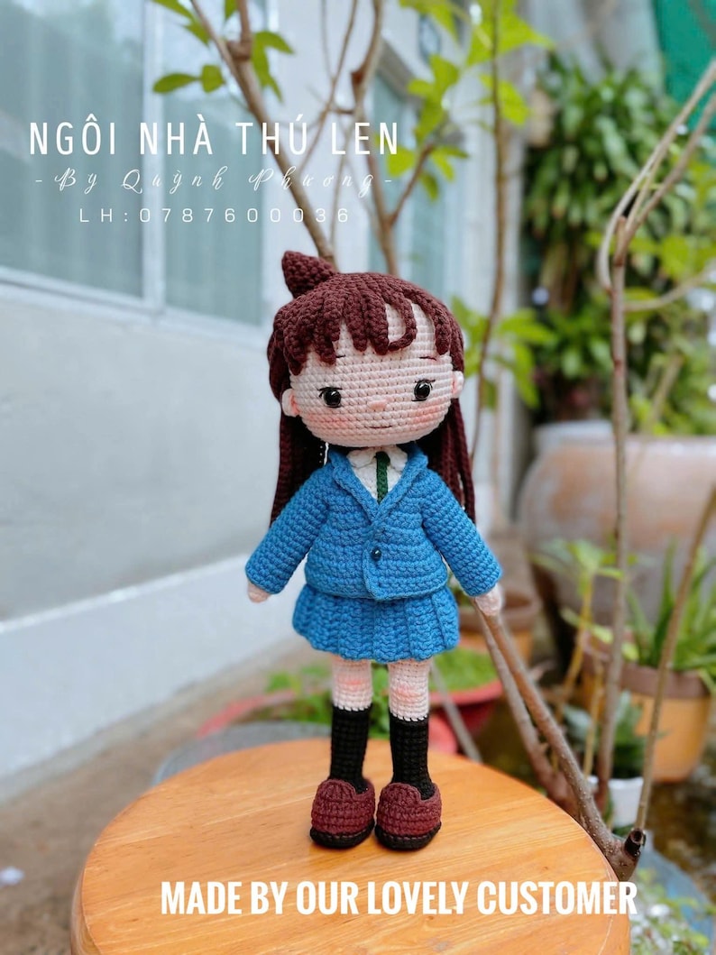 Pode incluir: Boneca de croch&ecirc; feita &agrave; m&atilde;o, vestindo uniforme escolar azul, saia combinando e sapatos bord&ocirc;. A boneca tem cabelo castanho e est&aacute; em uma superf&iacute;cie de madeira. Os textos "NG&Ocirc;I NH&Agrave; TH&Uacute; LEN" e "MADE BY OUR LOVELY CUSTOMER" s&atilde;o vis&iacute;veis.