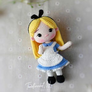 Puede incluir: Mu&ntilde;eca de ganchillo hecha a mano que representa a Alicia del pa&iacute;s de las maravillas. La mu&ntilde;eca tiene cabello amarillo, un lazo negro y viste un vestido azul y blanco. La cara de la mu&ntilde;eca tiene mejillas rosadas y ojos negros. La mu&ntilde;eca mide unos 15 cm de altura.