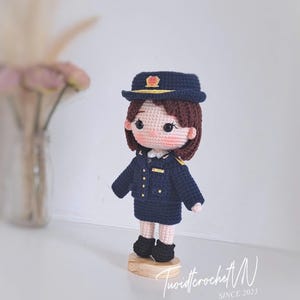 May include: A handcrafted crochet doll depicting a figure in a dark blue uniform, including a matching peaked cap with a gold emblem, a jacket with gold buttons, and a skirt. The doll has brown hair, rosy cheeks, and stands on a circular wooden base.