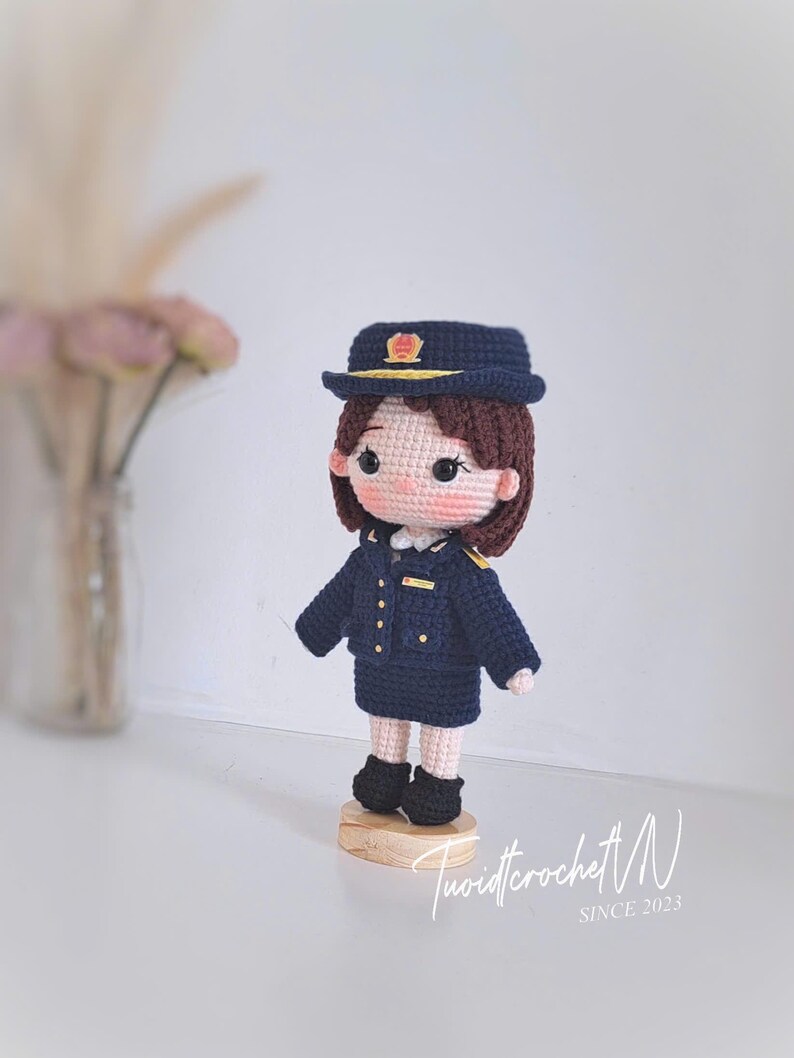 May include: A handcrafted crochet doll depicting a figure in a dark blue uniform, including a matching peaked cap with a gold emblem, a jacket with gold buttons, and a skirt. The doll has brown hair, rosy cheeks, and stands on a circular wooden base.