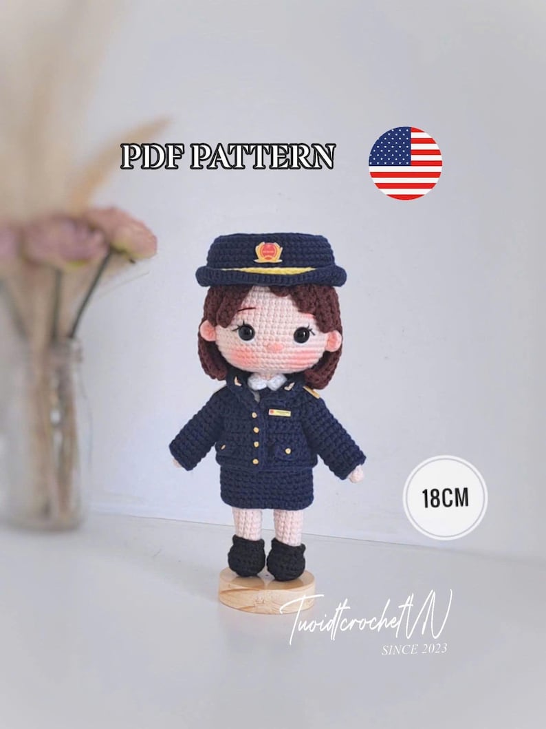 May include: A crochet doll wearing a dark blue uniform with a matching cap, featuring brown hair, black eyes, and pink cheeks. The doll stands on a wooden base and is labeled 18CM and PDF PATTERN with a US flag icon. The text TuoidtcrochetN SINCE 2023 is visible.