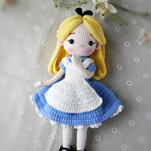 May include: Crocheted doll of a character with blonde hair, wearing a blue dress with a white apron and black shoes. The doll has a black bow and is set against a floral background. The doll is handmade.