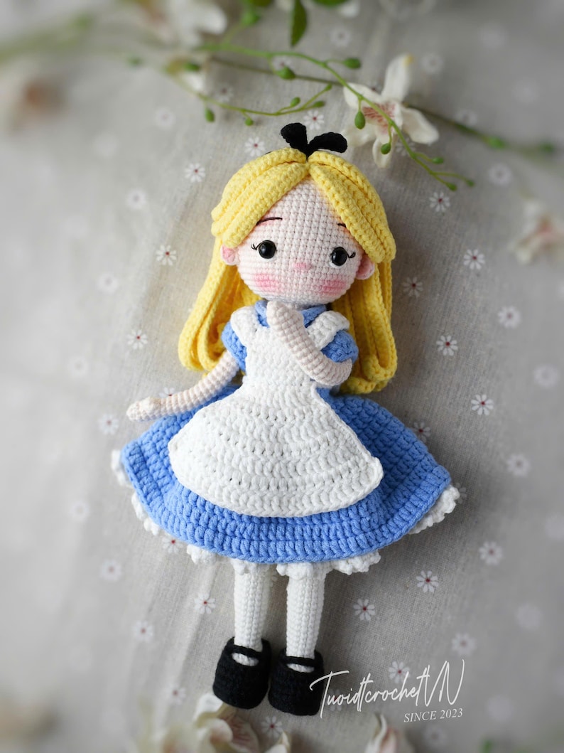May include: Crocheted doll of a character with blonde hair, wearing a blue dress with a white apron and black shoes. The doll has a black bow and is set against a floral background. The doll is handmade.