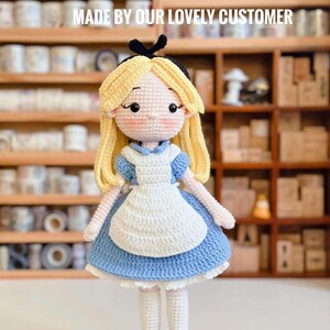 May include: A handmade crochet doll of Alice in Wonderland, featuring a blue dress, white apron, and blonde hair. The doll has black shoes and a black bow. The text "MADE BY OUR LOVELY CUSTOMER" is displayed above the doll.