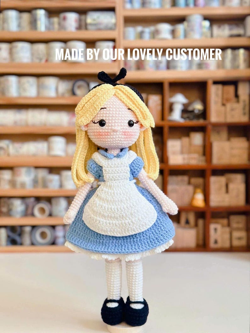May include: A handmade crochet doll of Alice in Wonderland, featuring a blue dress, white apron, and blonde hair. The doll has black shoes and a black bow. The text "MADE BY OUR LOVELY CUSTOMER" is displayed above the doll.