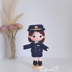 May include: A handcrafted crochet doll dressed in a dark navy blue uniform consisting of a jacket, skirt, and matching peaked cap with a yellow emblem. The doll has brown hair, black eyes, and stands on a small circular wooden base. The text TuoidicrochetN SINCE 2023 is visible.
