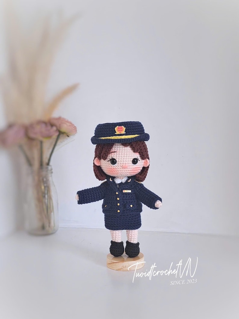 May include: A handcrafted crochet doll dressed in a dark navy blue uniform consisting of a jacket, skirt, and matching peaked cap with a yellow emblem. The doll has brown hair, black eyes, and stands on a small circular wooden base. The text TuoidicrochetN SINCE 2023 is visible.