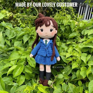 Pode incluir: Boneca de croch&ecirc; feita &agrave; m&atilde;o, com cabelo castanho, vestindo um casaco e saia azuis, uma camisa branca e botas pretas. A boneca tem tom de pele claro e est&aacute; rodeada por folhagem verde. O texto na imagem diz "MADE BY OUR LOVELY CUSTOMER" e "@yunmiu".
