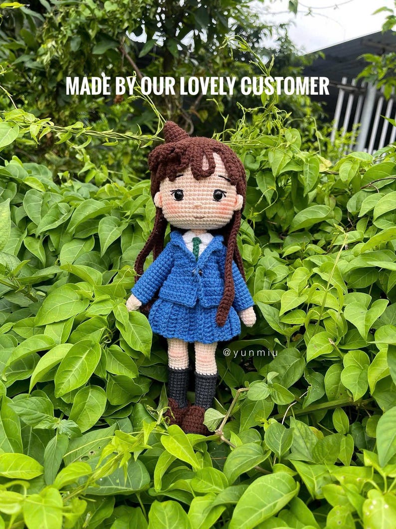 Pode incluir: Boneca de croch&ecirc; feita &agrave; m&atilde;o, com cabelo castanho, vestindo um casaco e saia azuis, uma camisa branca e botas pretas. A boneca tem tom de pele claro e est&aacute; rodeada por folhagem verde. O texto na imagem diz "MADE BY OUR LOVELY CUSTOMER" e "@yunmiu".