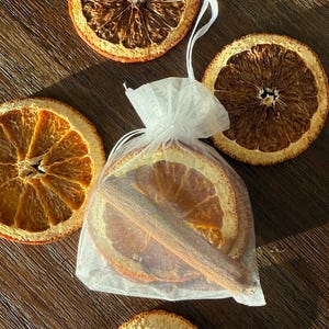 May include: A small white sheer organza drawstring bag containing dried orange slices and a cinnamon stick, surrounded by four additional dried orange slices arranged on a dark brown wooden surface.