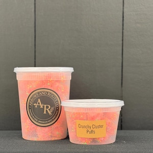 May include: Two clear plastic containers filled with colorful sprinkles. The larger container has a label with the text "Arrows and Roots Co." and a logo. The smaller container has a label that reads "Crunchy Cluster Puffs."