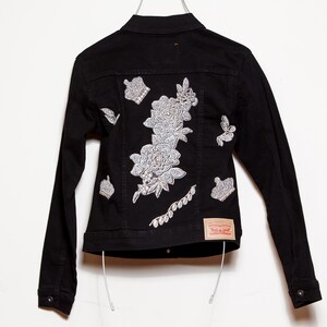 May include: Black denim jacket with white floral and crown embroidery on the back. The jacket has long sleeves, a button-down front, and a small red and white tag. A stylish piece of outerwear.