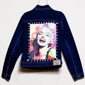 May include: Dark blue denim jacket featuring a colorful pop art image of Marilyn Monroe on the back. The image is framed with decorative stitching. The jacket has a classic design with button closures and pockets, perfect for casual wear.