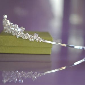 May include: A silver tiara adorned with clear gemstones, displayed on a small, olive green box. The tiara has a design of teardrop and round-cut stones. The background is a blurred purple, with the tiara's reflection visible.