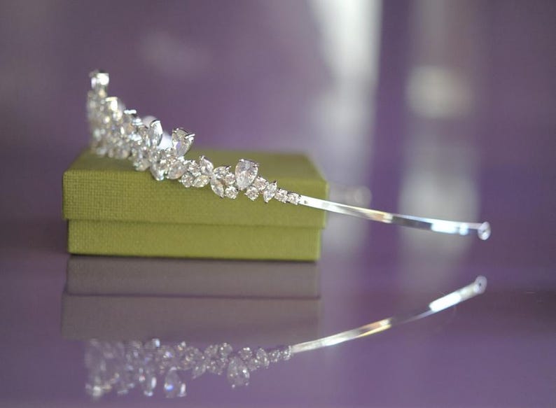 May include: A silver tiara adorned with clear gemstones, displayed on a small, olive green box. The tiara has a design of teardrop and round-cut stones. The background is a blurred purple, with the tiara's reflection visible.