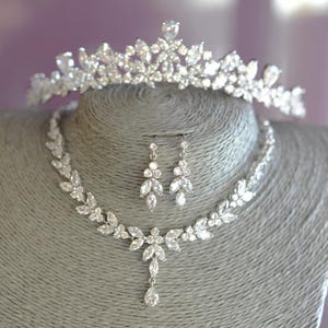 May include: A silver-toned bridal jewellery set comprising a tiara, necklace, and earrings, all embellished with clear crystals. The necklace features a leaf design and a teardrop pendant. The set is displayed on a grey mannequin.
